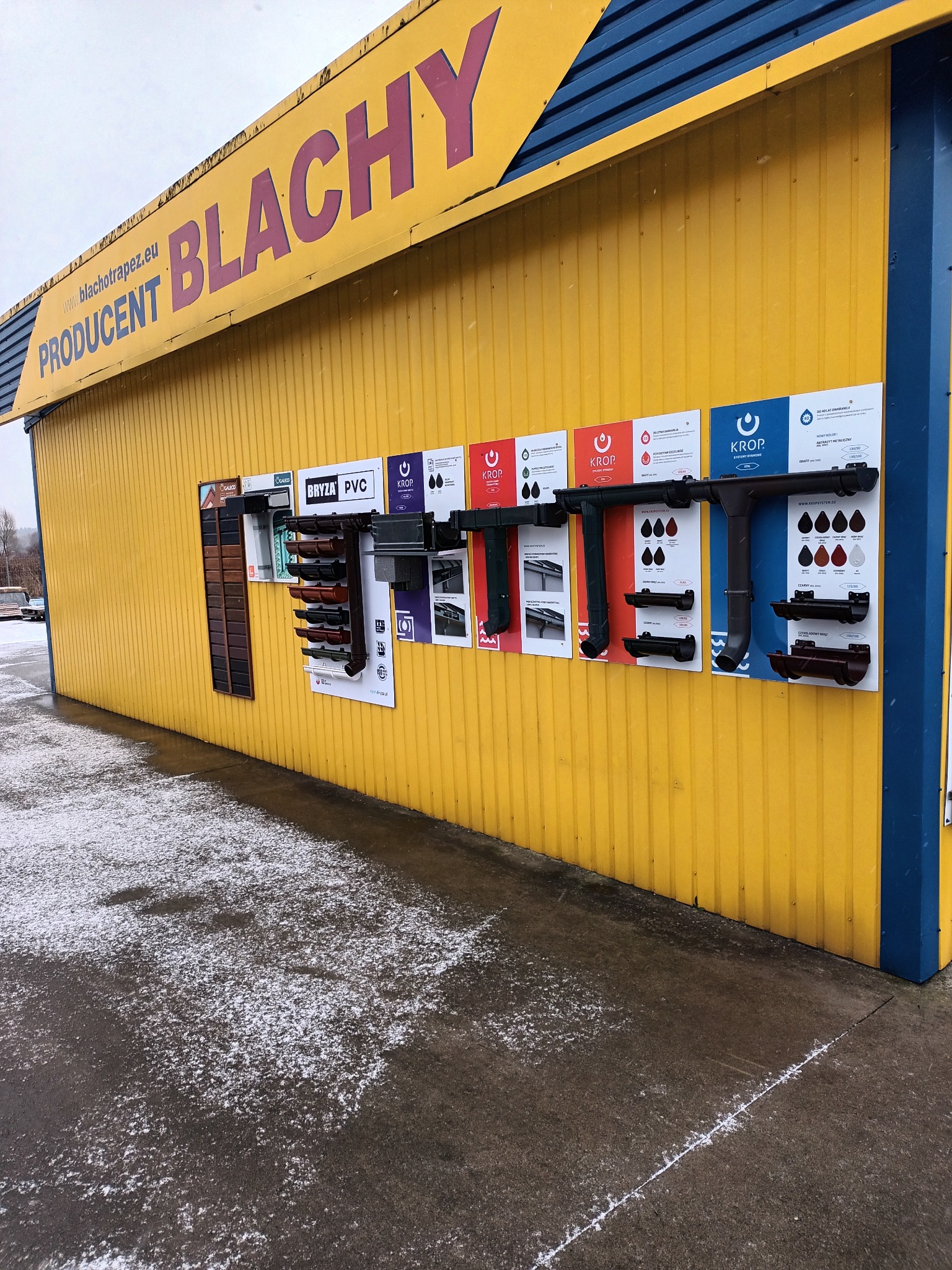 Ekspozycja rynien różnych typów i kolorów na ścianie budynku składu budowlanego z żółtą elewacją i napisem 'BLACHY'.