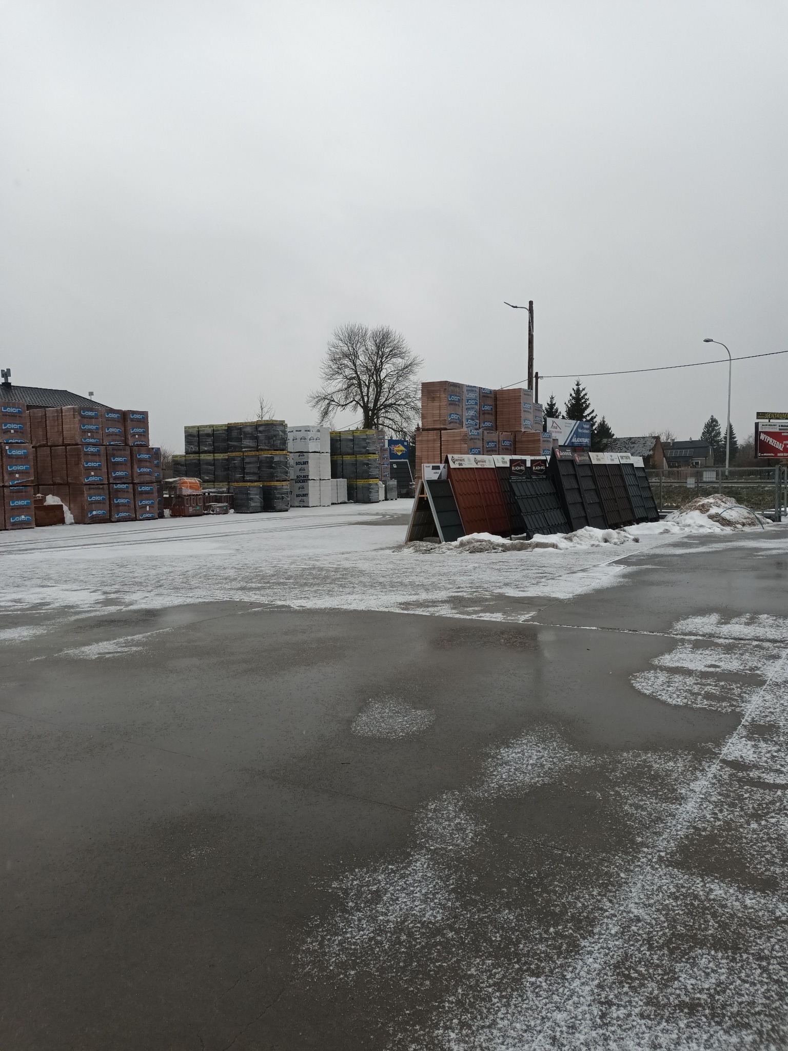 Składowisko materiałów budowlanych z paletami cegieł i dachówek na ekspozytorach, lekko przyprószone śniegiem, pochmurny dzień.