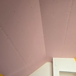 Uslugi remontovo - budowlane - Montaż ognioodpornych płyt gipsowych na poddaszu w Pucku. Widoczne łączenia płyt i wkręty. Żółta taśma zabezpieczająca na styku ściany i sufitu.