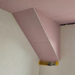 Uslugi remontovo - budowlane - Różowa płyta karton-gips zamontowana na suficie i ścianie, tworząca skośną konstrukcję. Widoczne ślady po wkrętach i fragment żółtej izolacji. W tle białe ściany.