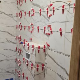 Uslugi remontovo - budowlane - Łazienka w trakcie remontu: białe płytki imitujące marmur na ścianach, czerwone klipsy poziomujące, drewniana boazeria na skosie sufitu. Widoczne narzędzia.