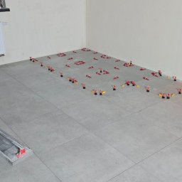 Uslugi remontovo - budowlane - Szare płytki podłogowe w trakcie układania, z systemem poziomowania (czerwone kliny i czarne podstawki). W rogu pomieszczenia widoczny fragment narzędzia do cięcia płytek.
