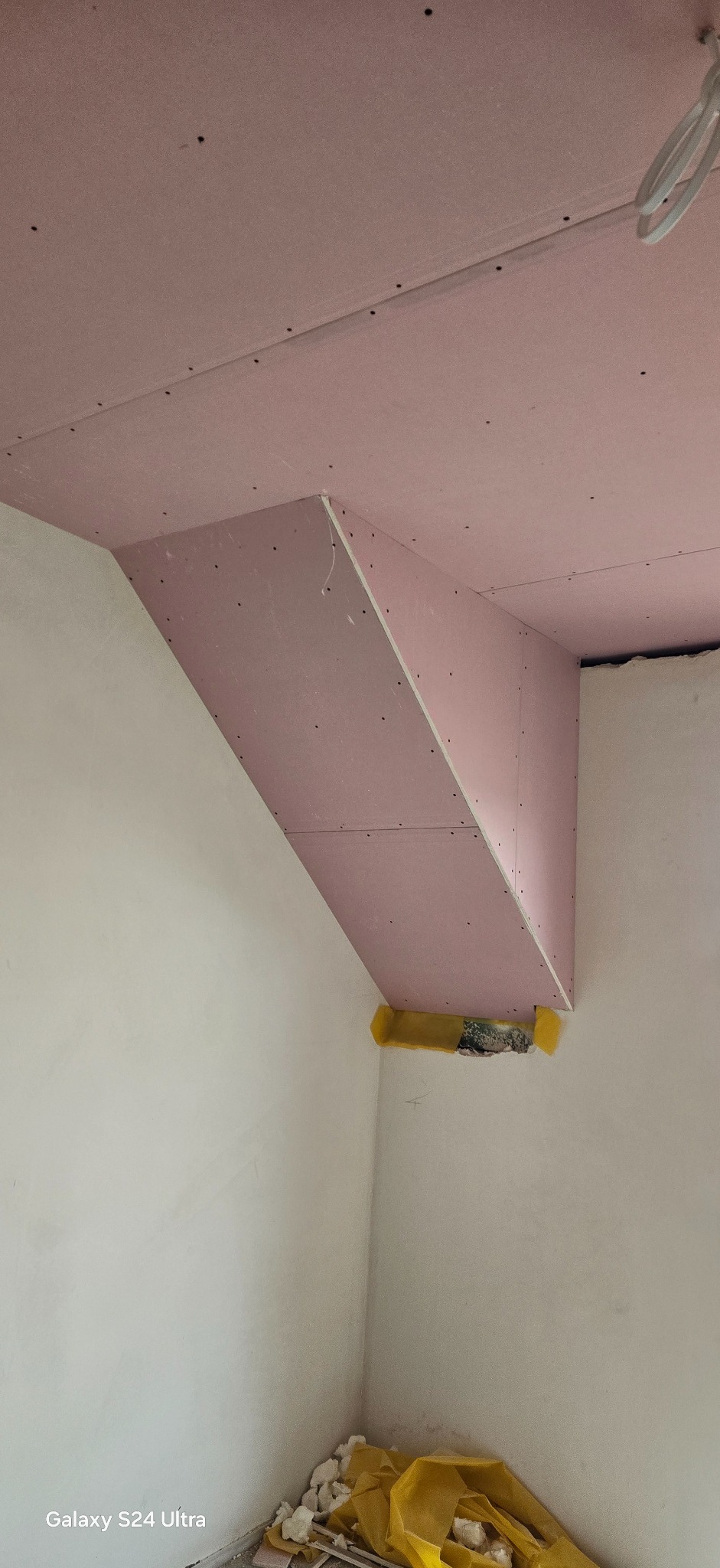 Różowa płyta karton-gips zamontowana na suficie i ścianie, tworząca skośną konstrukcję. Widoczne ślady po wkrętach i fragment żółtej izolacji. W tle białe ściany.