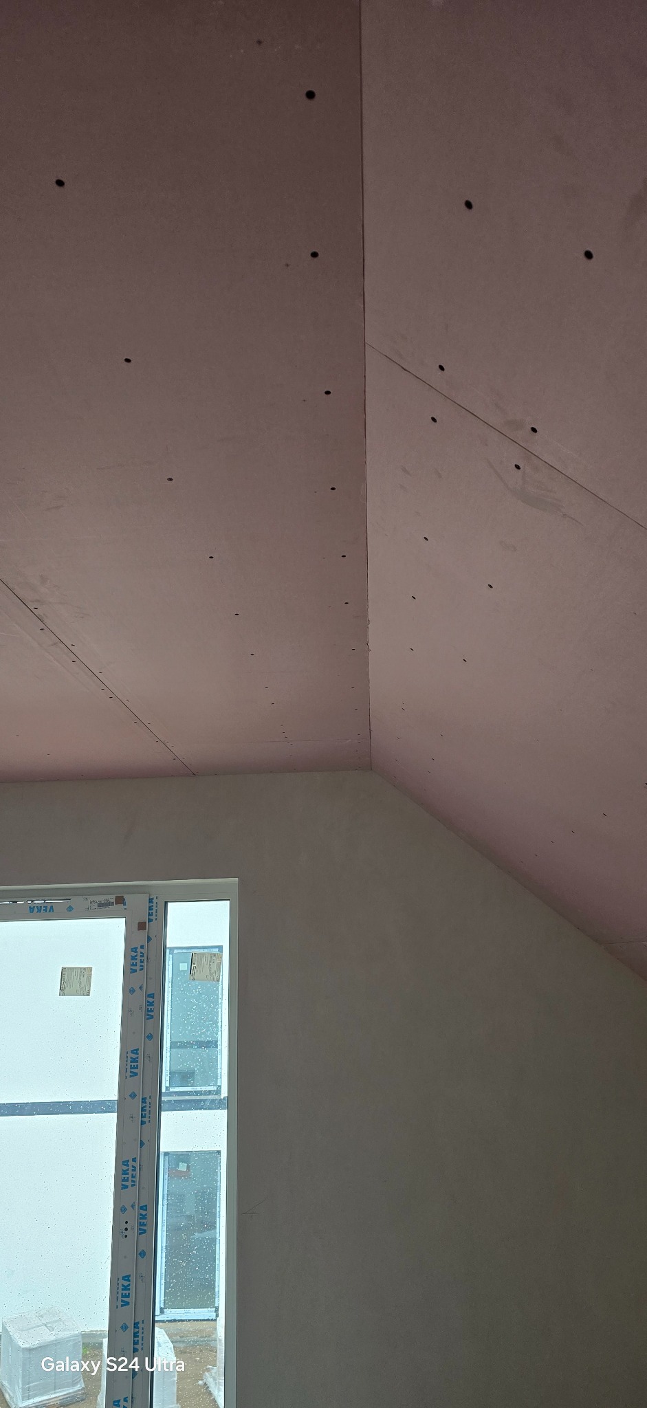 Skośny sufit poddasza z płyt kartonowo-gipsowych, widoczne wkręty, okno z białymi ramami. Ściana szpachlowana, przygotowana do malowania.