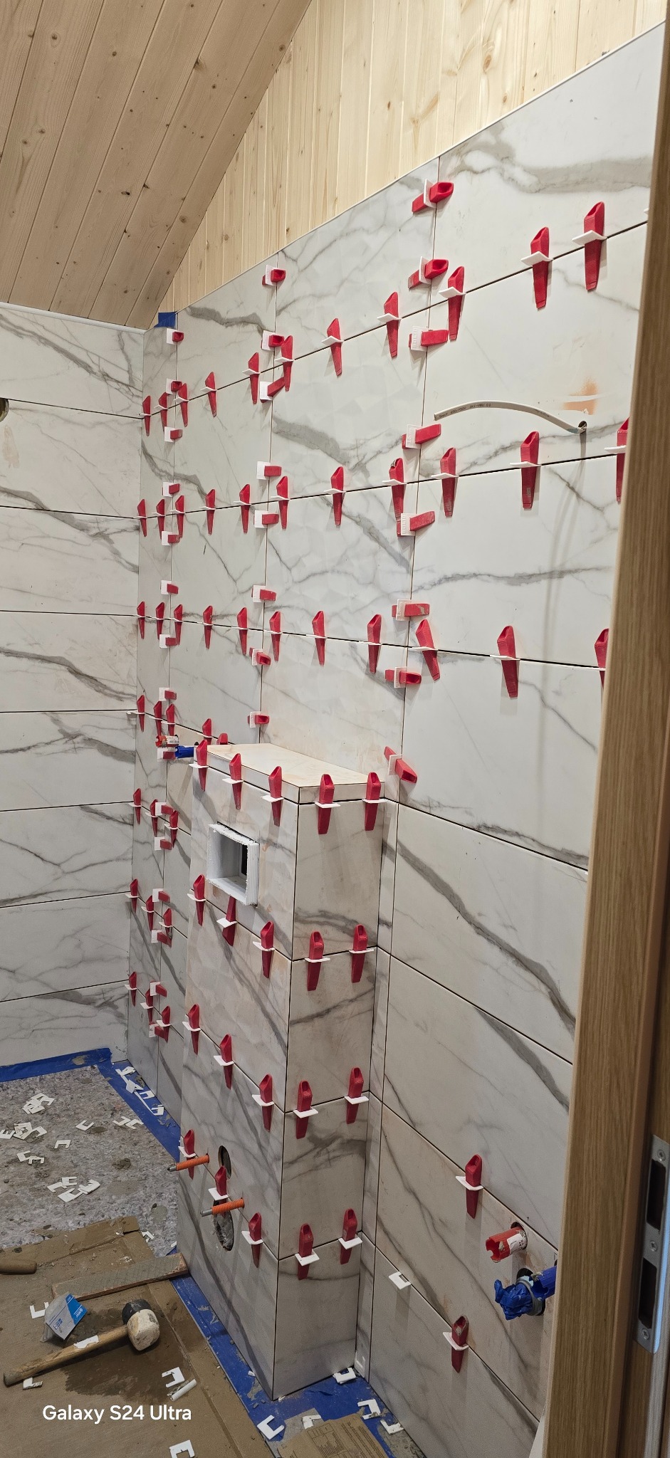 Łazienka w trakcie remontu: białe płytki imitujące marmur na ścianach, czerwone klipsy poziomujące, drewniana boazeria na skosie sufitu. Widoczne narzędzia.