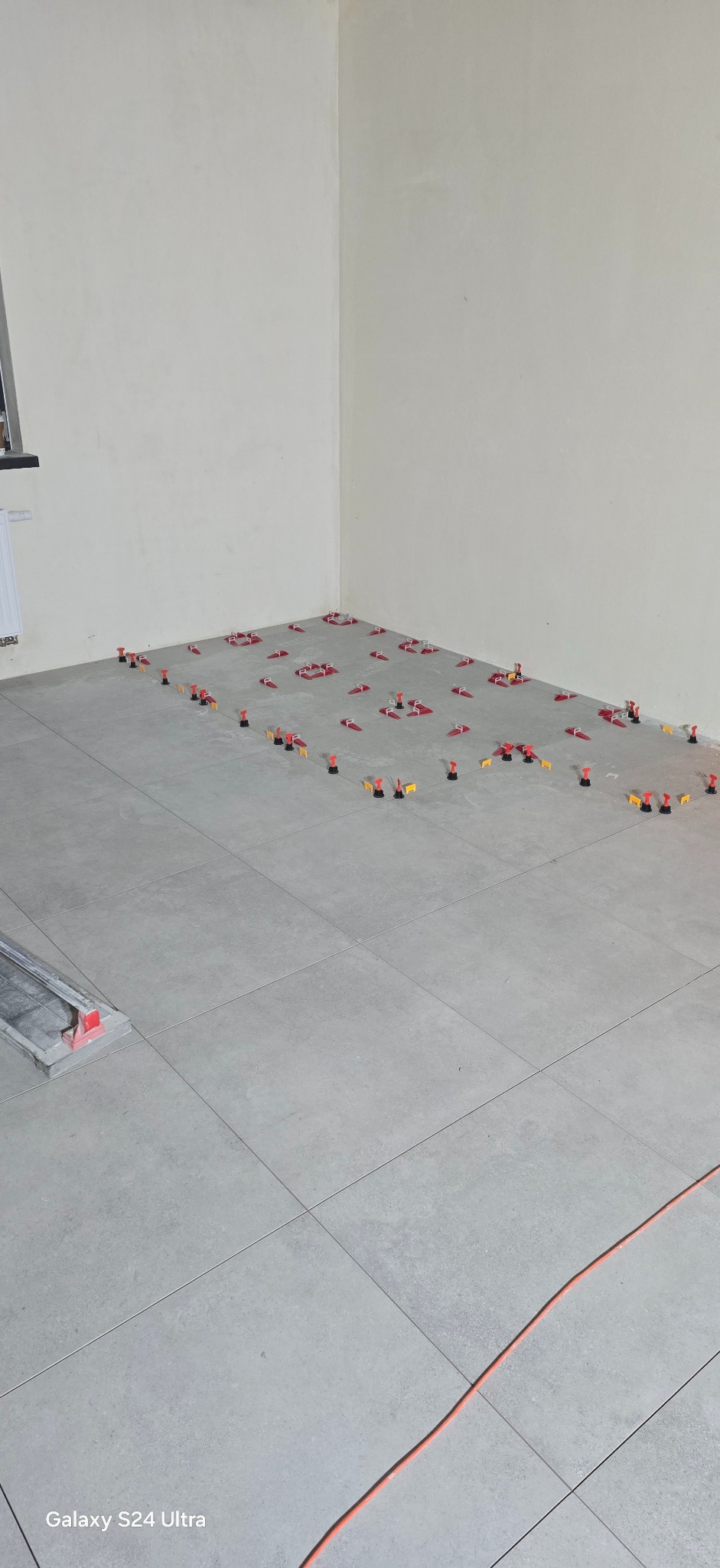 Szare płytki podłogowe w trakcie układania, z systemem poziomowania (czerwone kliny i czarne podstawki). W rogu pomieszczenia widoczny fragment narzędzia do cięcia płytek.