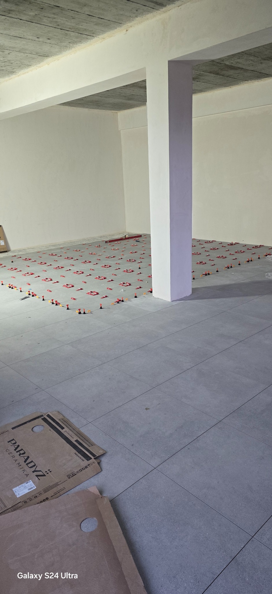 Układanie szarych płytek podłogowych w przestronnym wnętrzu z użyciem poziomujących klipsów i klinów. Kartony płytek Paradysz w rogu. Widoczny betonowy sufit.