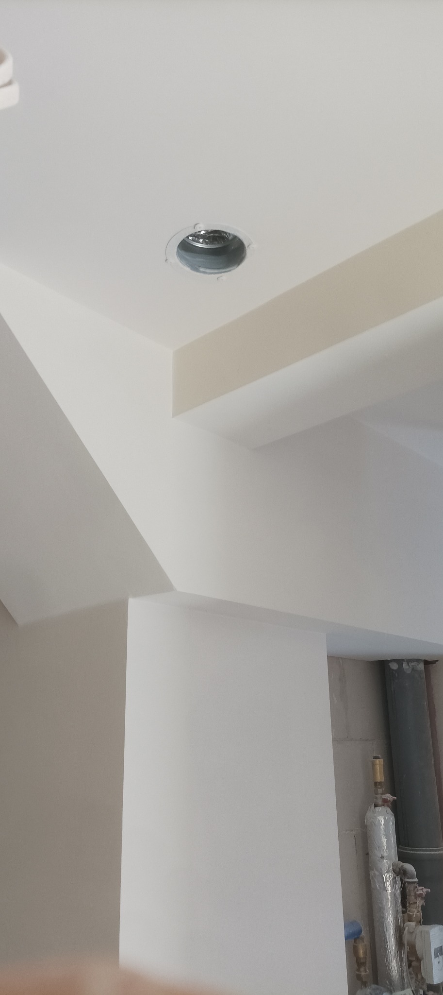 Białe, świeżo pomalowane wnętrze z widocznym otworem wentylacyjnym w suficie i fragmentem instalacji. Minimalistyczny design, jasne barwy, surowe wykończenie.