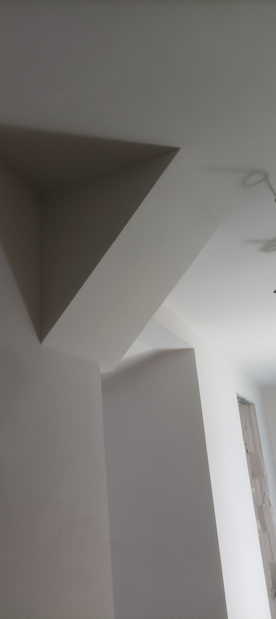 Białe ściany i sufit z nietypową, trójkątną wnęką. Widoczny fragment okna w stanie surowym. Minimalistyczne wykończenie wnętrza.