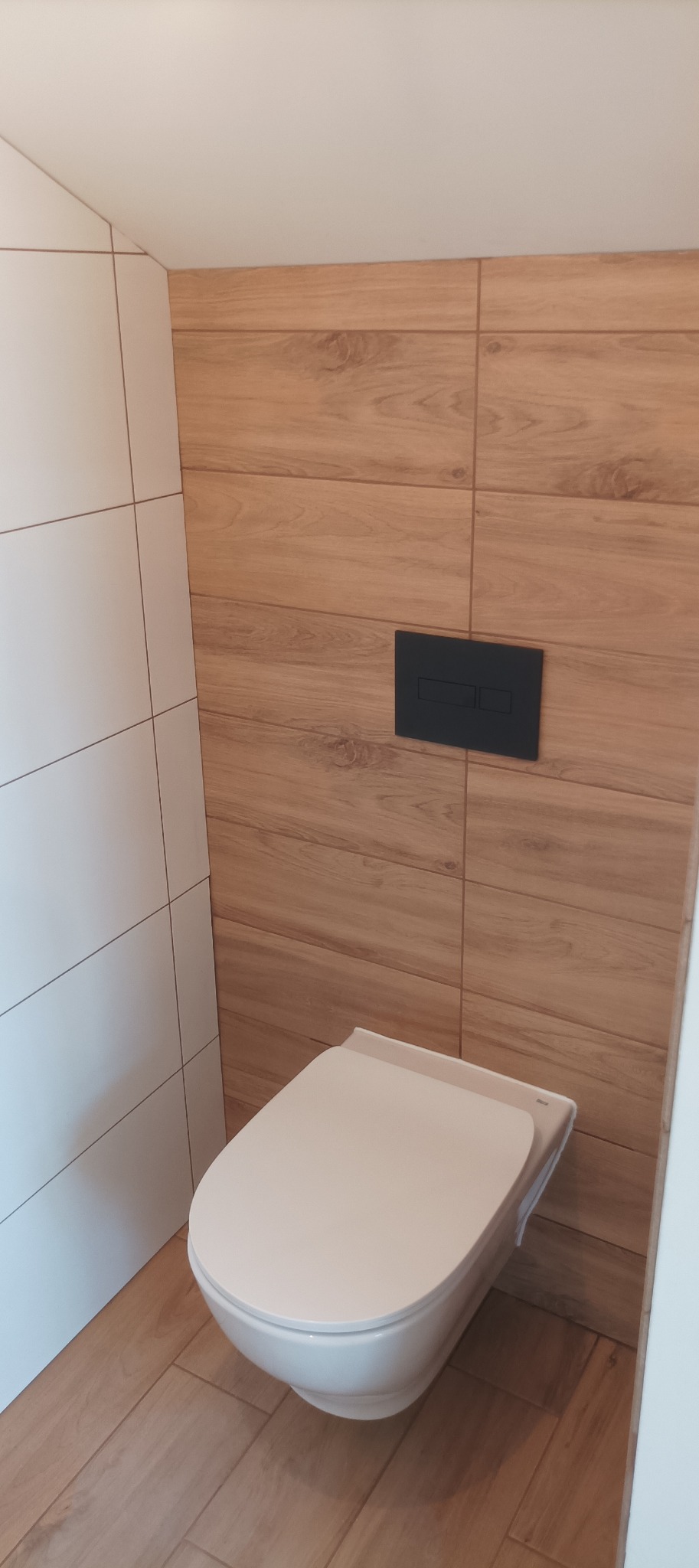 Nowoczesna toaleta podwieszana z czarnym przyciskiem spłukującym na ścianie wyłożonej płytkami imitującymi drewno. Minimalistyczny design łazienki.