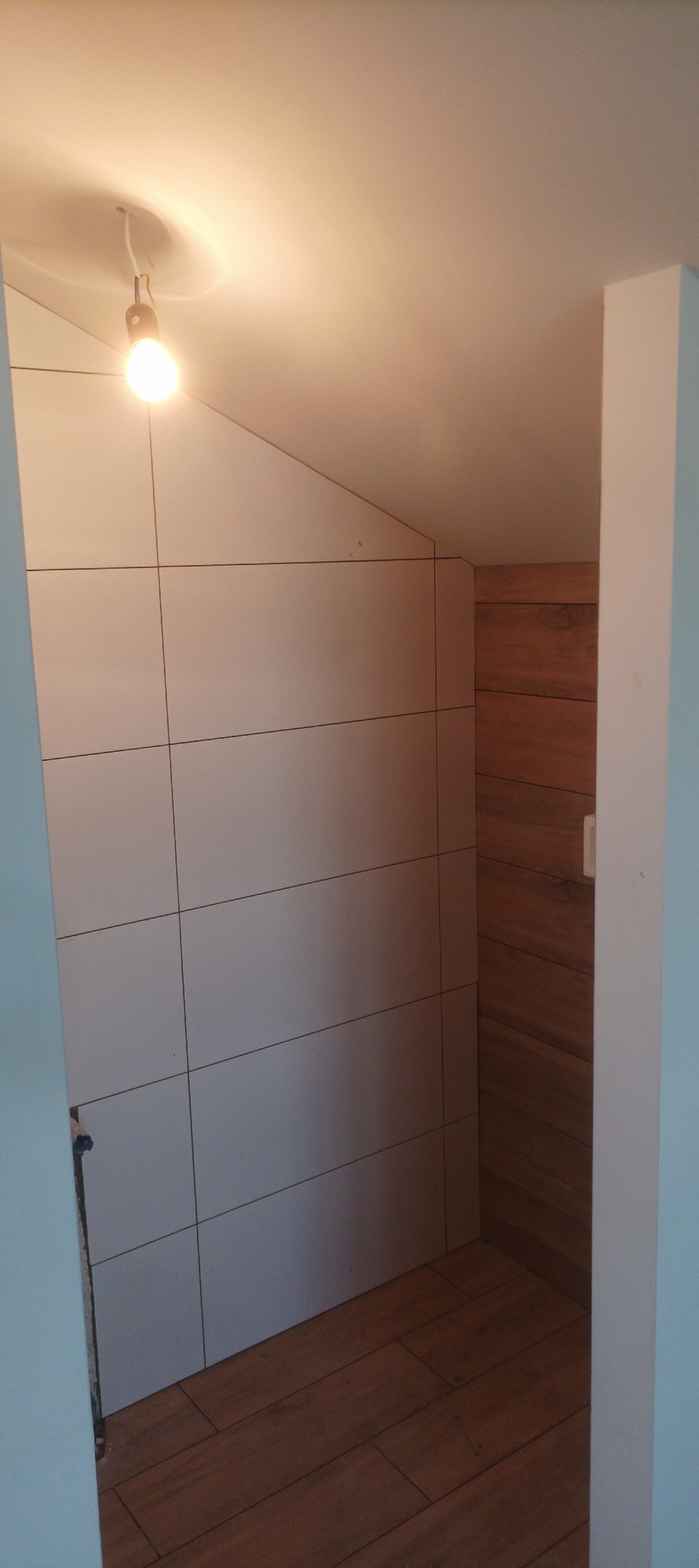 Wykończona łazienka na poddaszu z białymi płytkami ściennymi i drewnopodobną podłogą. Ukośna ściana z płytkami, ciepłe oświetlenie. Widok z wnętrza pomieszczenia.