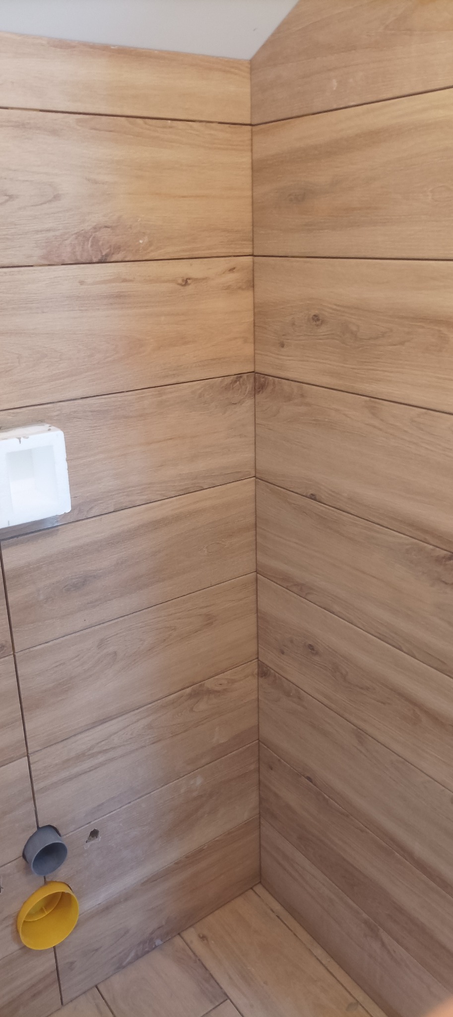 Wykończona ściana w łazience z drewnopodobnych płytek, widoczne otwory na instalacje. Ułożenie płytek w jodełkę. Jasne, ciepłe oświetlenie.