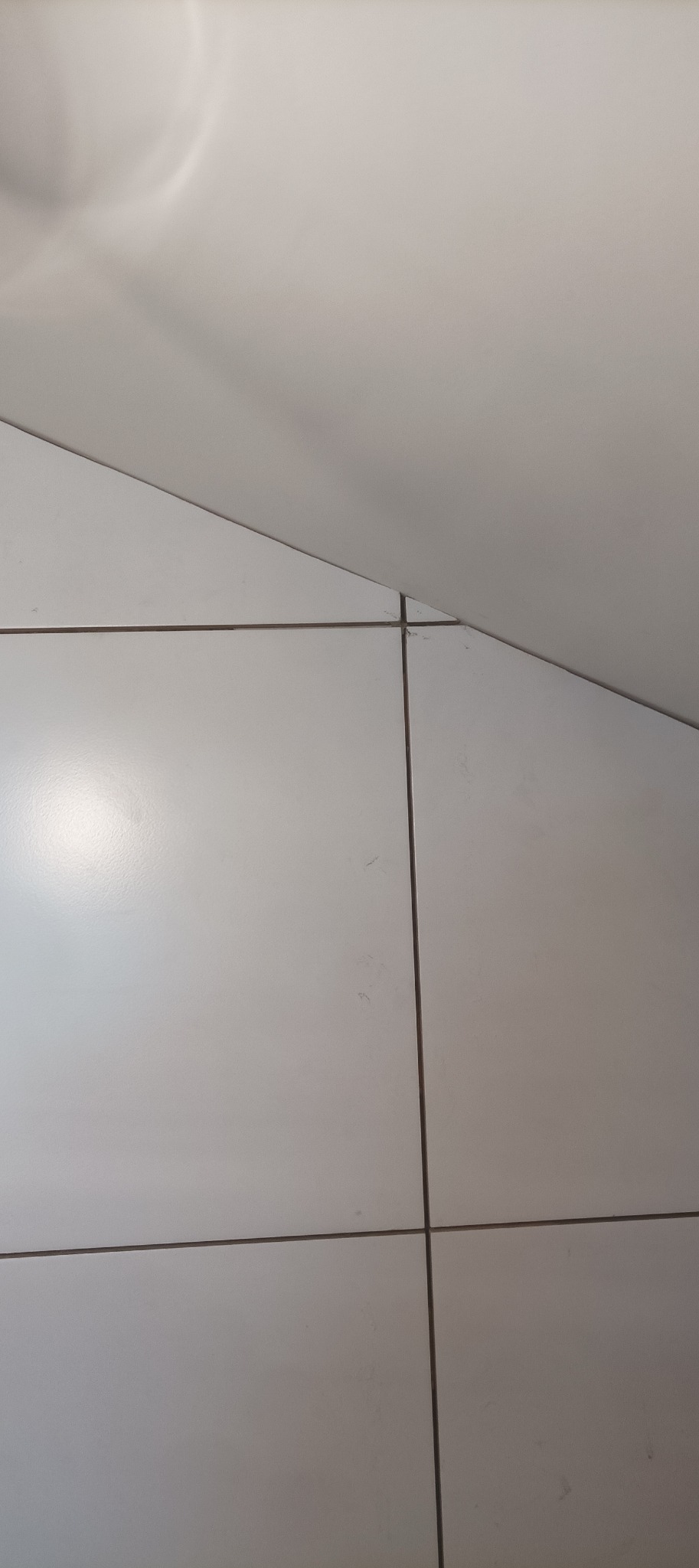 Białe, kwadratowe płytki ceramiczne ułożone na podłodze lub ścianie, z widocznymi fugami. Górny róg kadru zajmuje fragment białej, gładkiej powierzchni, prawdopodobnie sufitu.