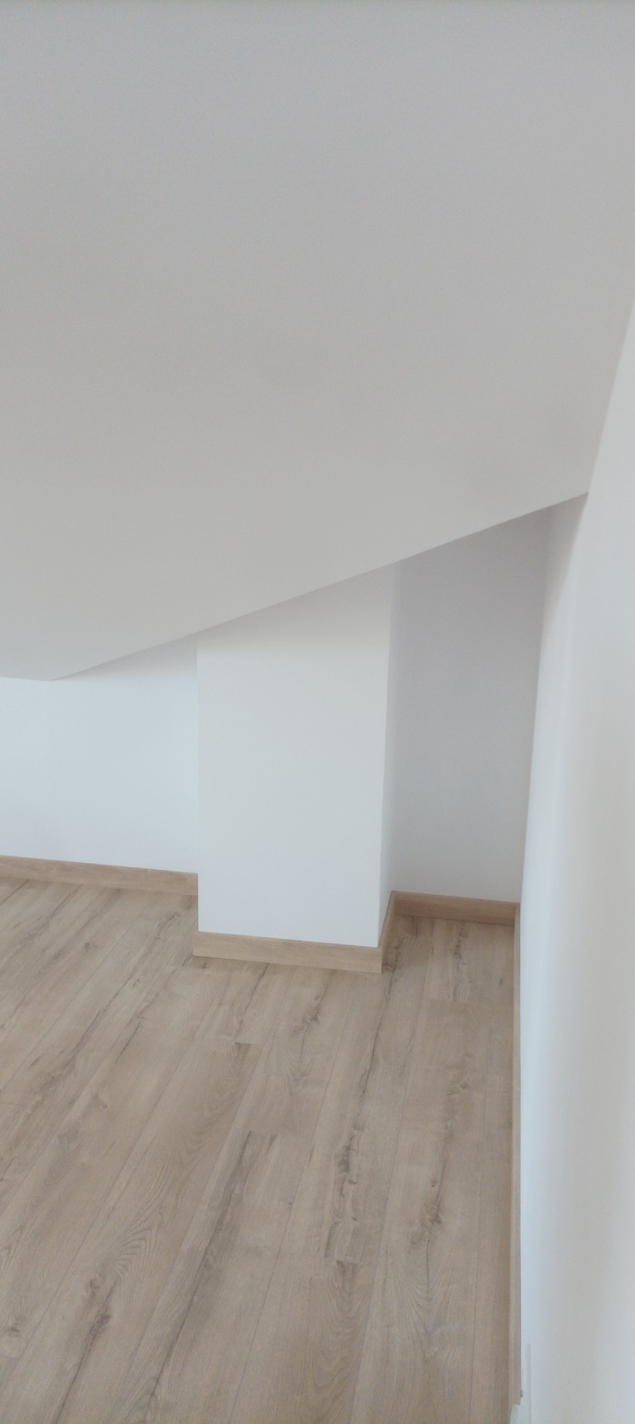 Jasne panele podłogowe w nowym, minimalistycznym wnętrzu z białymi ścianami i skośnym sufitem. Widoczne listwy przypodłogowe w kolorze drewna.