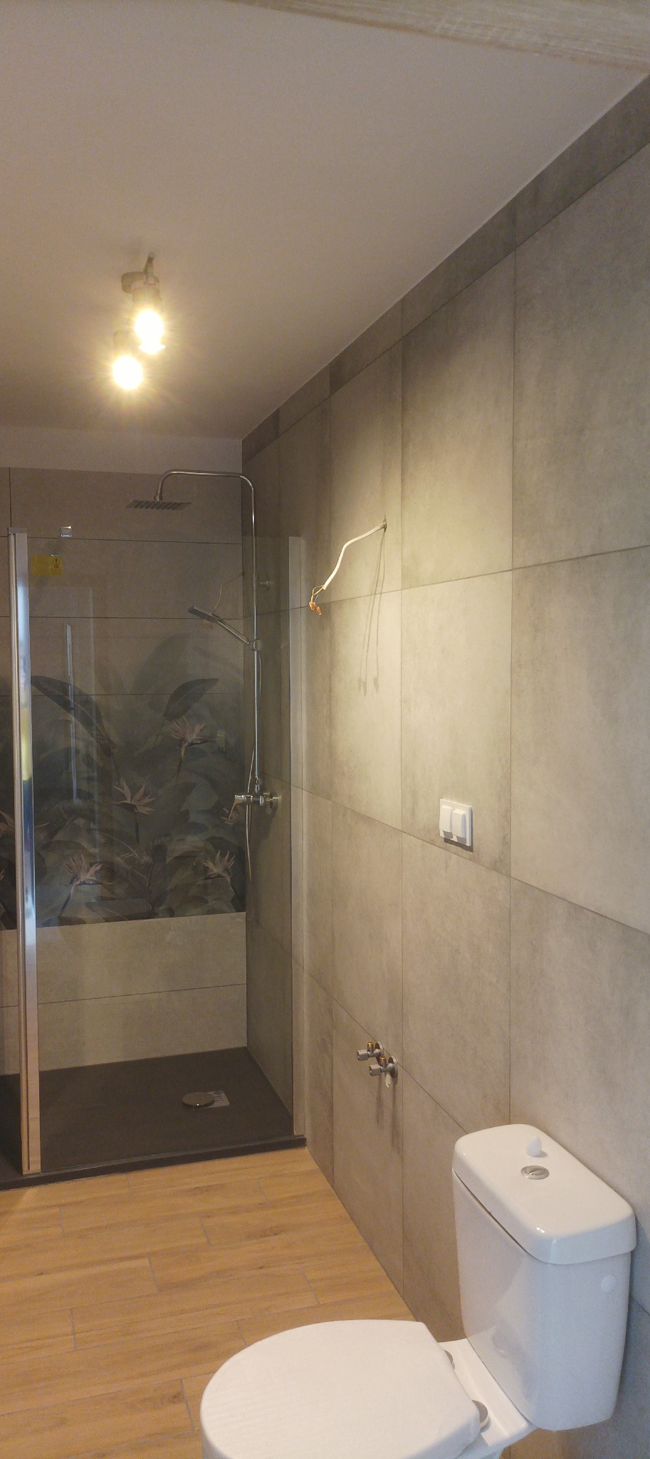 Nowoczesna łazienka z kabiną prysznicową typu walk-in, szarymi płytkami i drewnopodobną podłogą. Widoczne niedokończone podłączenie elektryczne.