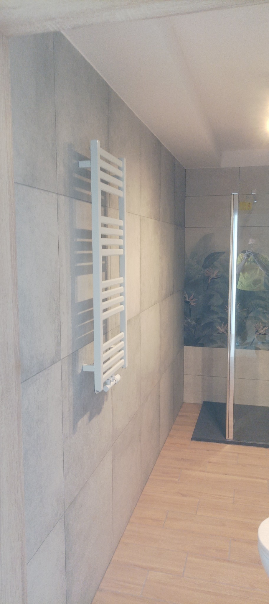 Wykończona łazienka z grzejnikiem drabinkowym na szarej, kwadratowej ścianie z płytek. Kabina prysznicowa z motywem roślinnym w tle. Podłoga wyłożona panelami.