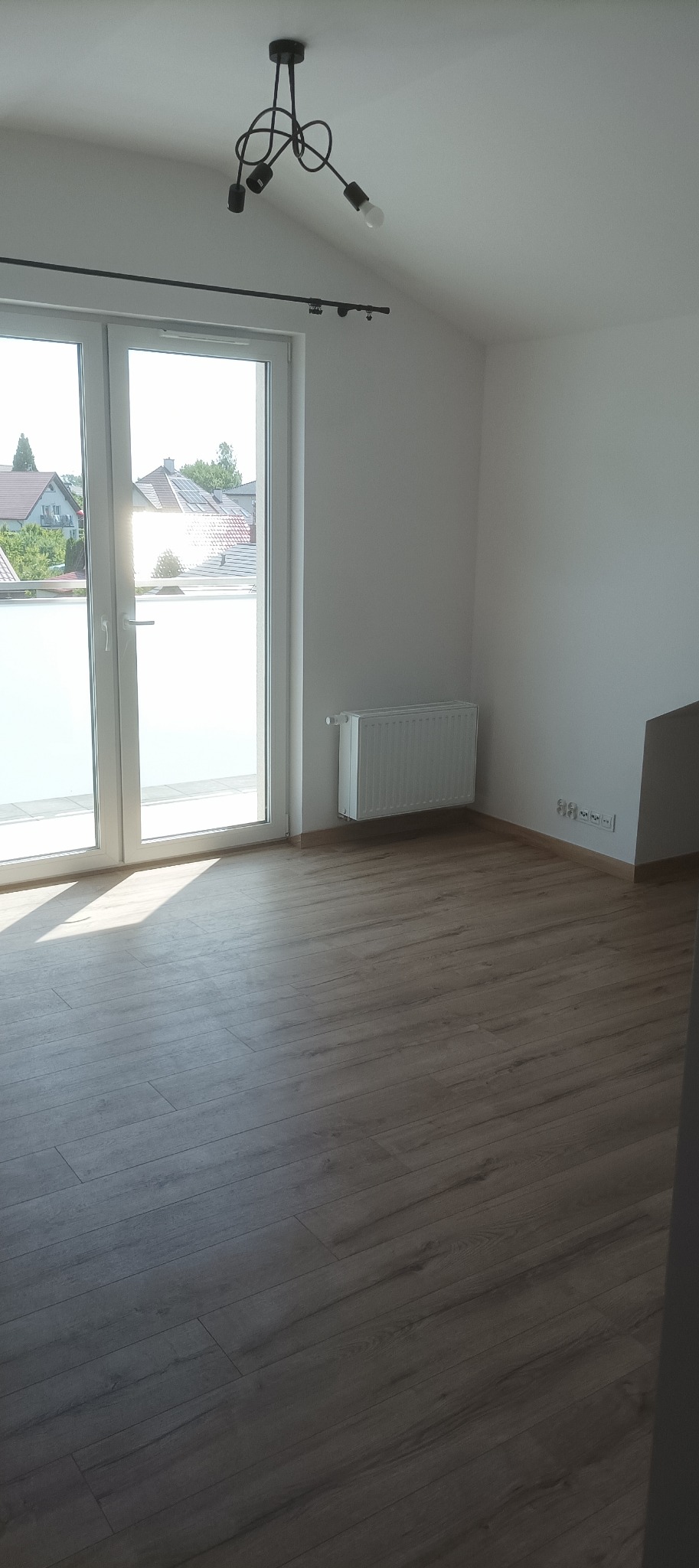 Jasne wnętrze po remoncie: panele podłogowe, białe ściany, grzejnik, okno balkonowe z widokiem na zewnątrz i nowoczesna lampa sufitowa.