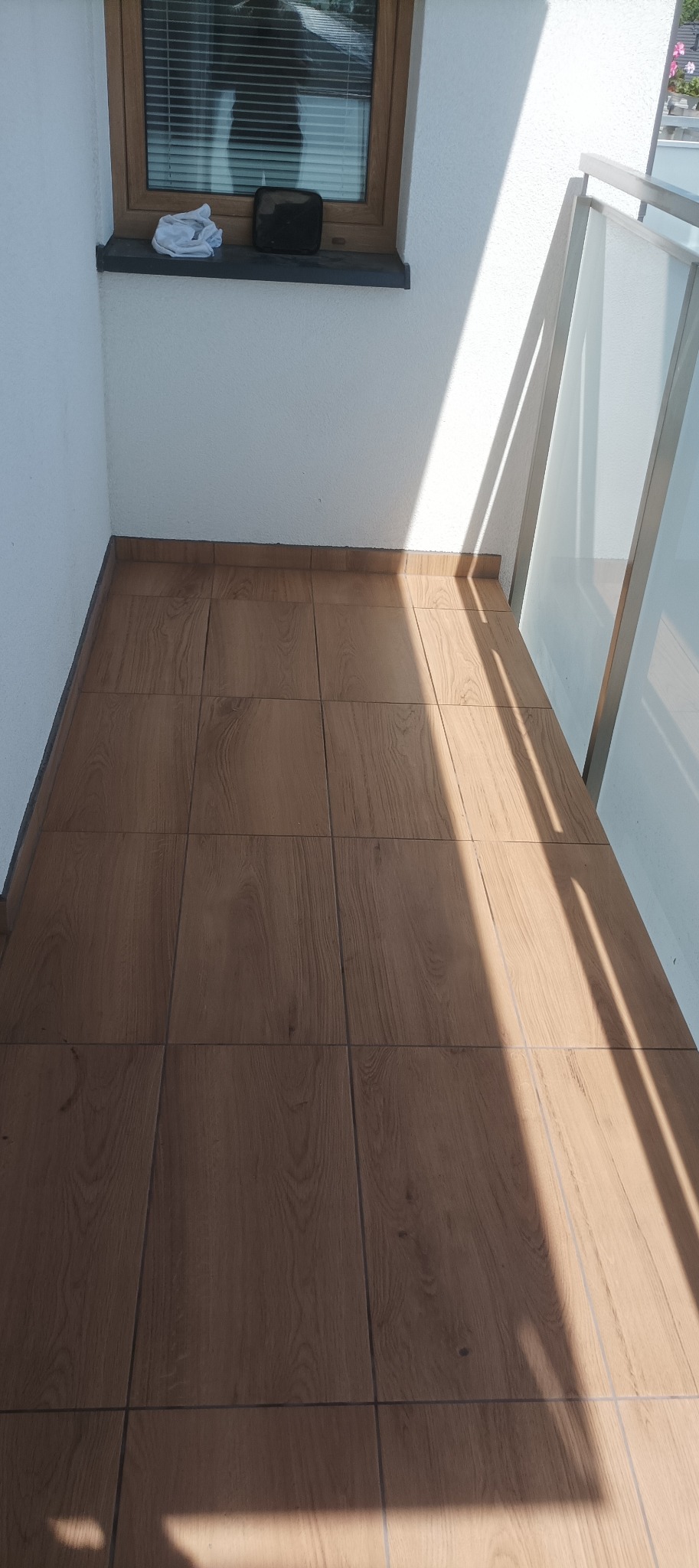 Balkon z drewnopodobną terakotą w ciepłym odcieniu, oświetlony promieniami słońca. Widoczne okno z żaluzjami i fragment balustrady z mlecznego szkła.