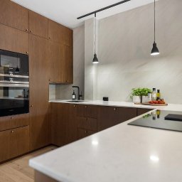 Szafa Na Miare Grzegorz Tyndyk - Kuchnia w stylu modern classic z drewnianymi frontami, wbudowanym piekarnikiem Siemens, białym blatem i czarną armaturą, oświetlona wiszącymi lampami i dekorowana ziołami w doniczkach.