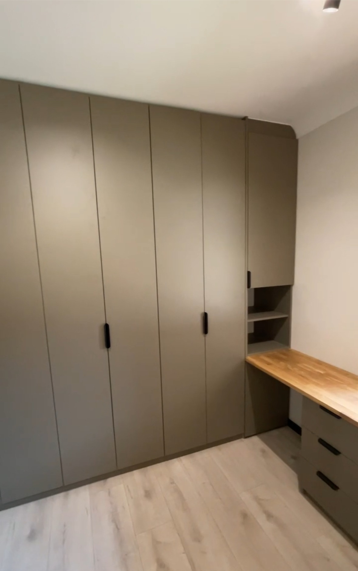 Wbudowana szafa z szarymi frontami i czarnymi uchwytami, połączona z biurkiem z drewnianym blatem i szufladami, na tle jasnej podłogi.