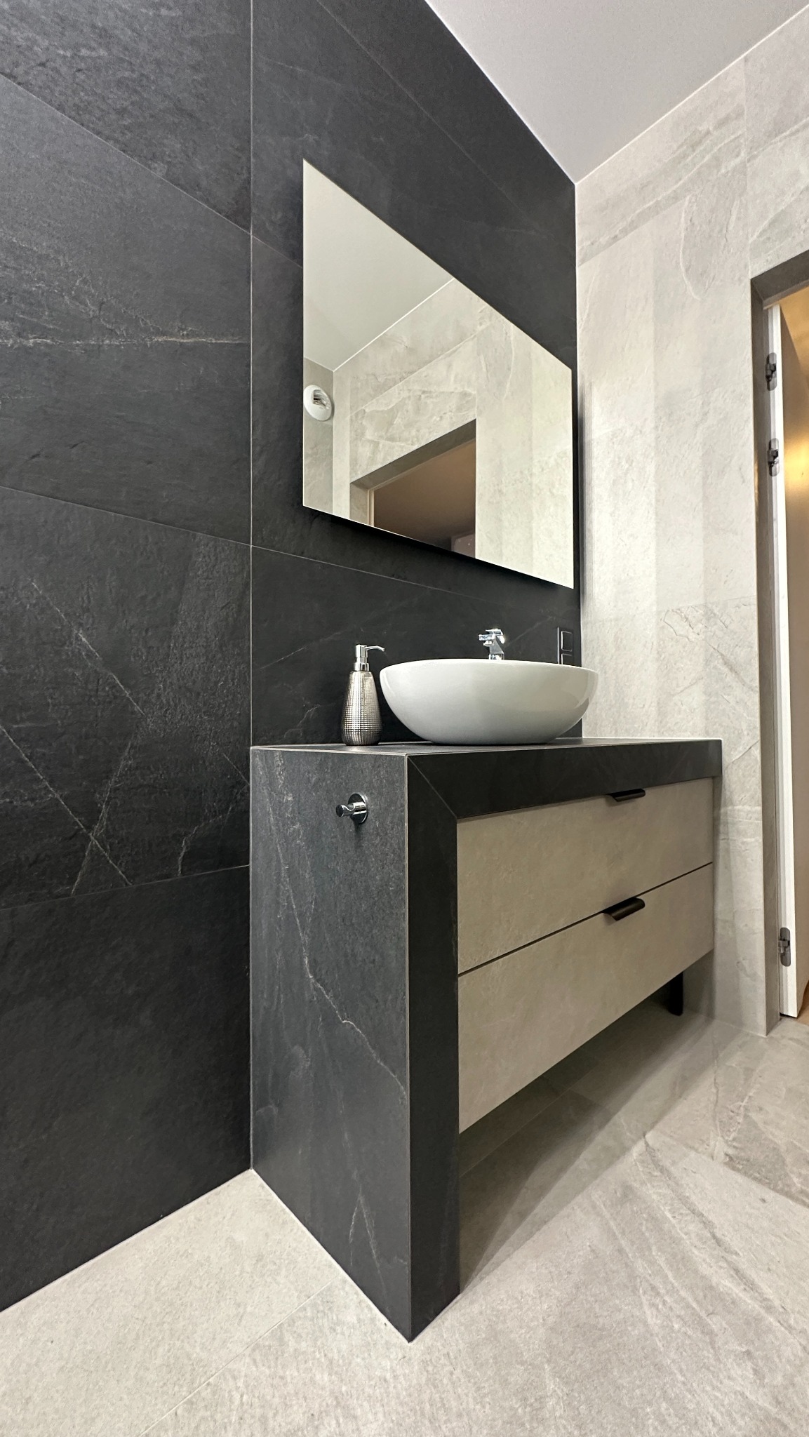 Nowoczesna łazienka z umywalką nablatową, szafką z szufladami w kolorze beżowym i czarnymi płytkami imitującymi marmur na ścianach, z lustrem w prostokątnej ramie.