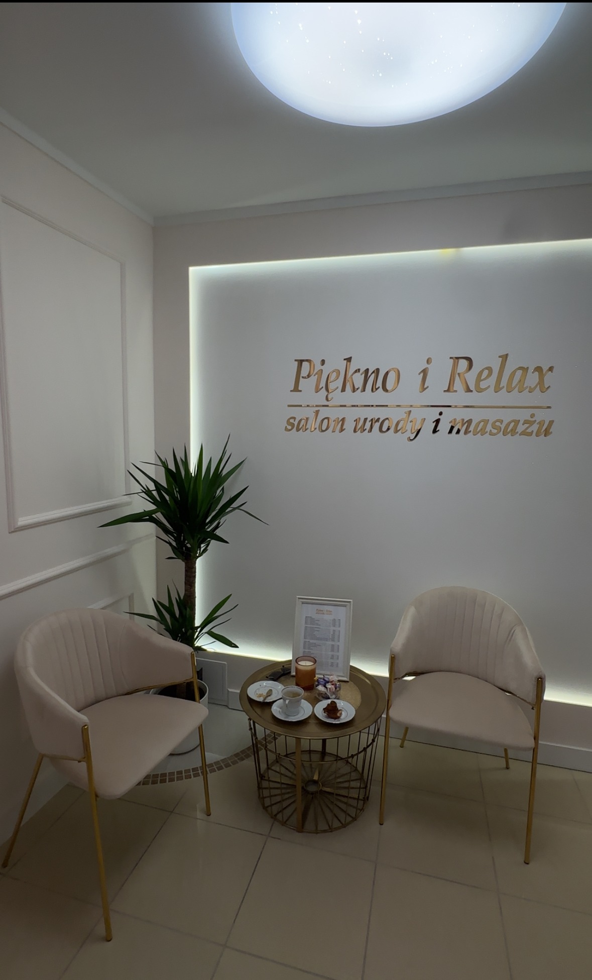 Jasne wnętrze salonu urody z dwoma fotelami, stolikiem kawowym z filiżankami i przekąskami, oraz rośliną w tle, podświetlone napisem 'Piękno i Relax salon urody i masażu'.