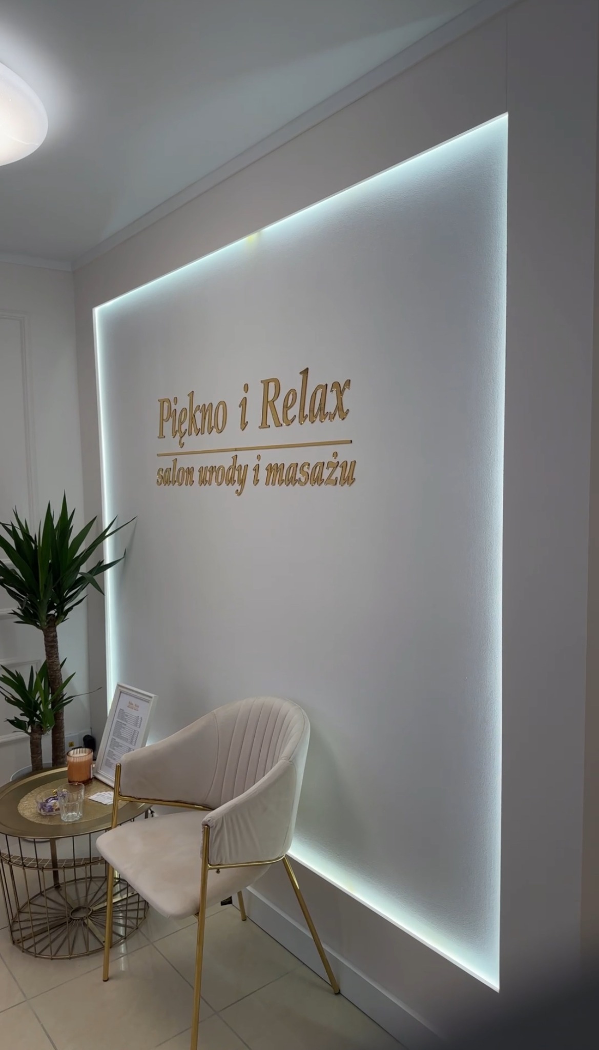 Recepcja salonu piękności: białe ściany z podświetleniem LED, złoty napis 'Piękno i Relax salon urody i masażu', beżowy fotel z metalowymi nogami i stolik z rośliną doniczkową.