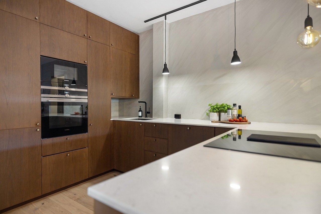 Kuchnia w stylu modern classic z drewnianymi frontami, wbudowanym piekarnikiem Siemens, białym blatem i czarną armaturą, oświetlona wiszącymi lampami i dekorowana ziołami w doniczkach.