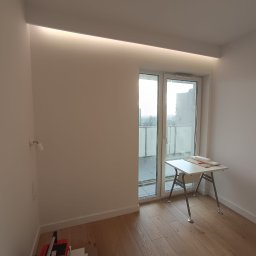 UNIPROFGROUP SPÓŁKA Z OGRANICZONĄ ODPOWIEDZIALNOŚCIĄ - Jasne wnętrze po remoncie: drewniana podłoga, białe ściany z oświetleniem LED, okno balkonowe i narzędzia do układania płytek w rogu. Minimalistyczny design.