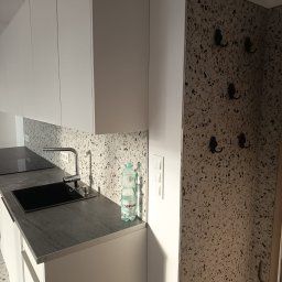 UNIPROFGROUP SPÓŁKA Z OGRANICZONĄ ODPOWIEDZIALNOŚCIĄ - Nowoczesna kuchnia z białymi szafkami, blatem z ciemnego kamienia i mozaikową ścianą. Bateria kuchenna i butelka wody na blacie. Haczyki na ścianie w tle.