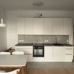 UNIPROFGROUP SPÓŁKA Z OGRANICZONĄ ODPOWIEDZIALNOŚCIĄ - Nowoczesna, minimalistyczna kuchnia z białymi szafkami, szarym blatem i mozaiką na ścianie. Widoczny stół z krzesłami i lampa wisząca. Uwagę zwraca wzór lastryko na podłodze.