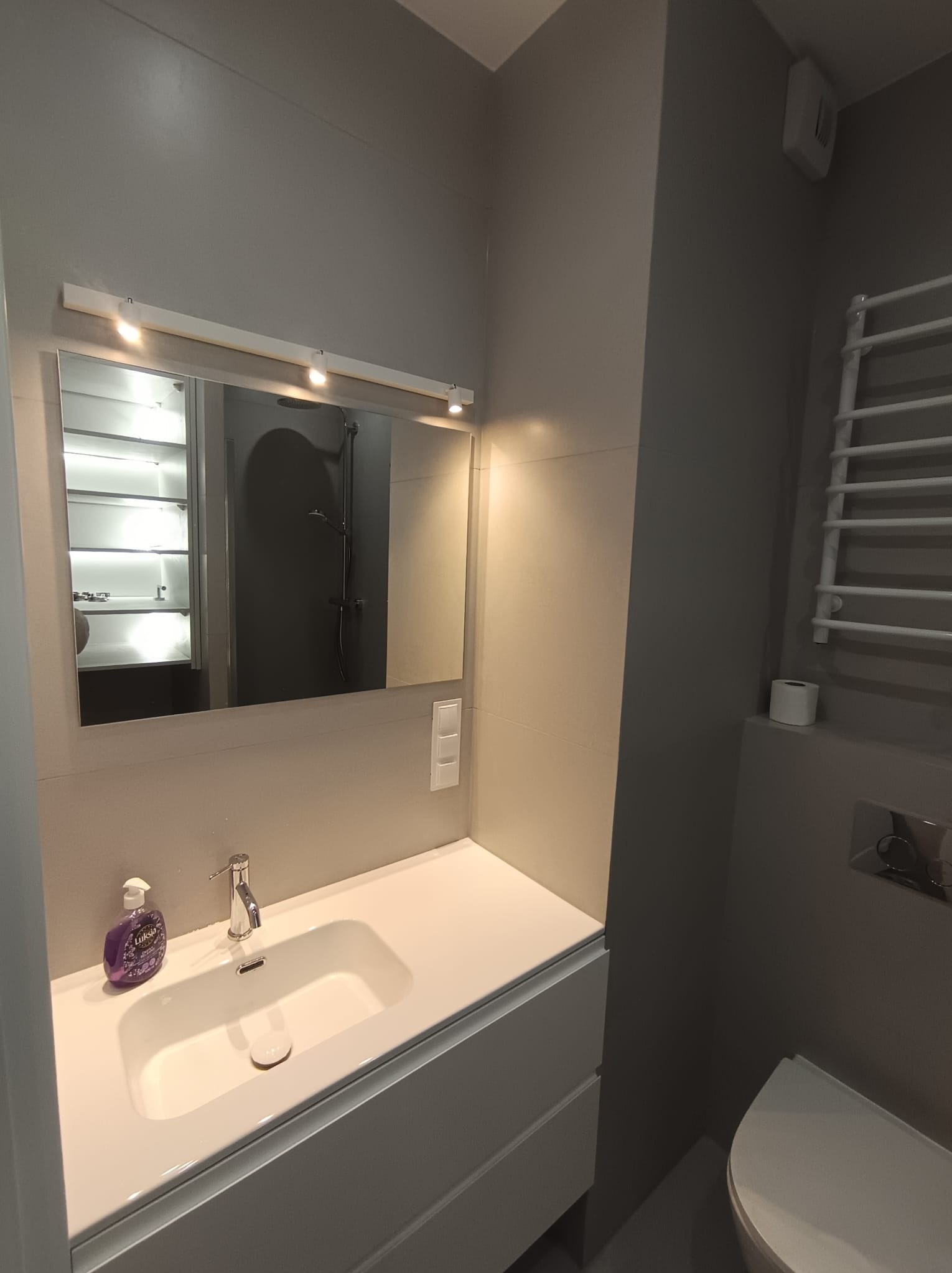 Nowoczesna łazienka w szarościach: biała umywalka z szafką, lustro z oświetleniem LED, grzejnik drabinkowy i toaleta. Minimalistyczny design, czyste linie.