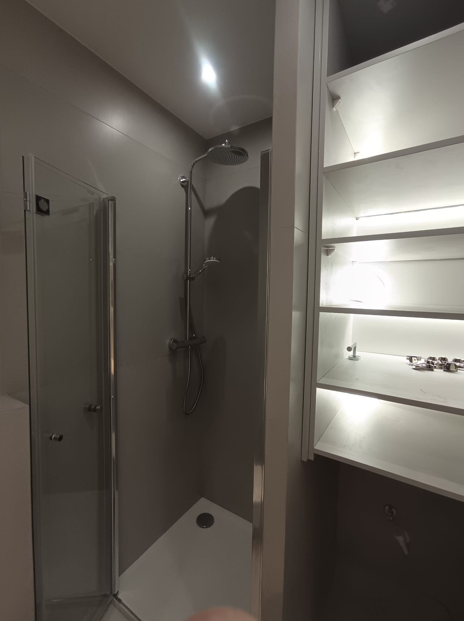 Nowoczesna kabina prysznicowa z deszczownicą i szklanymi drzwiami, obok białe półki z oświetleniem LED. Minimalistyczny design łazienki w odcieniach szarości i bieli.