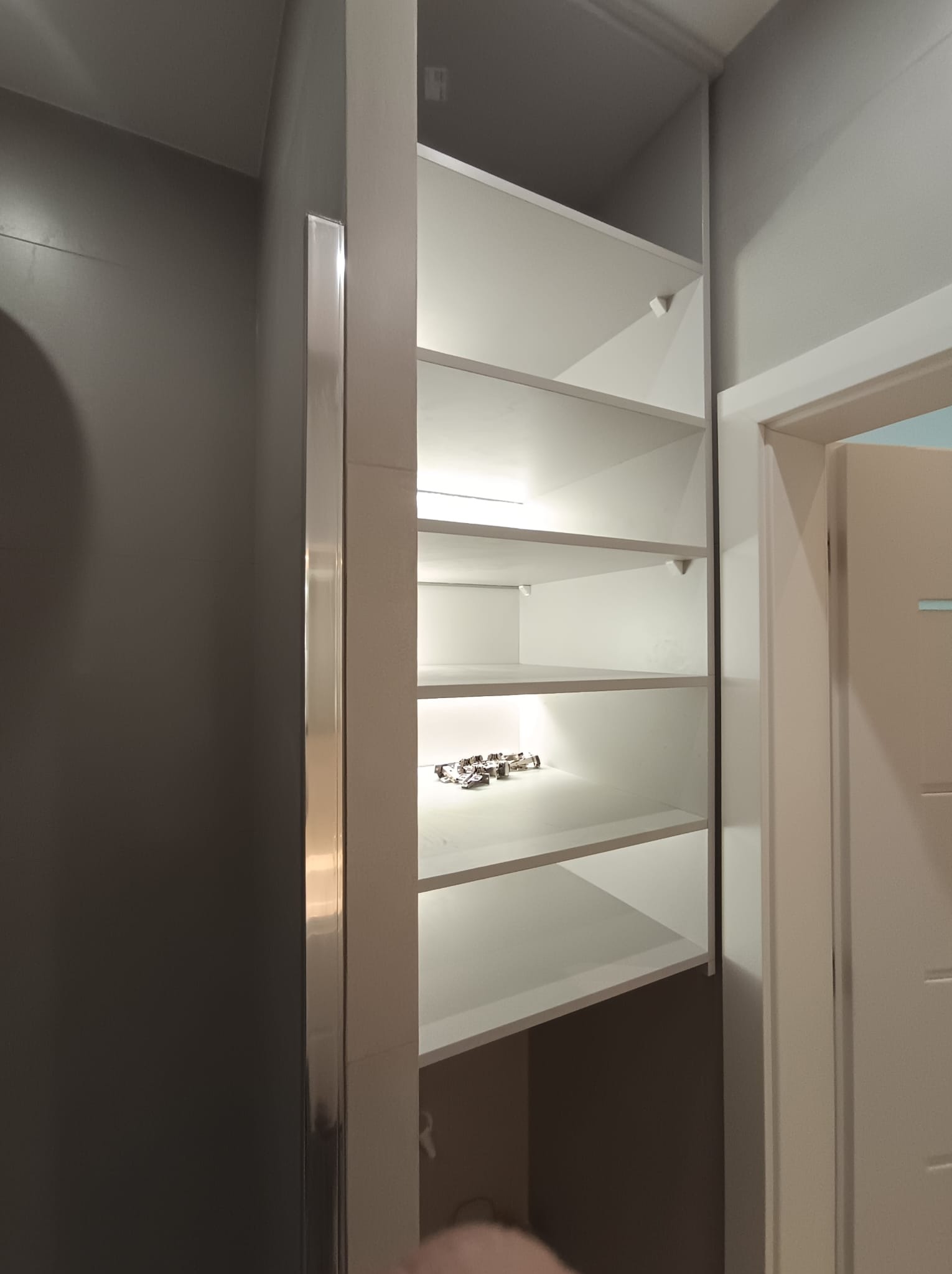 Wnęka z półkami w łazience, oświetlona od wewnątrz, z widocznymi zawiasami na jednej z półek. Szara ściana kontrastuje z bielą półek i drzwi.