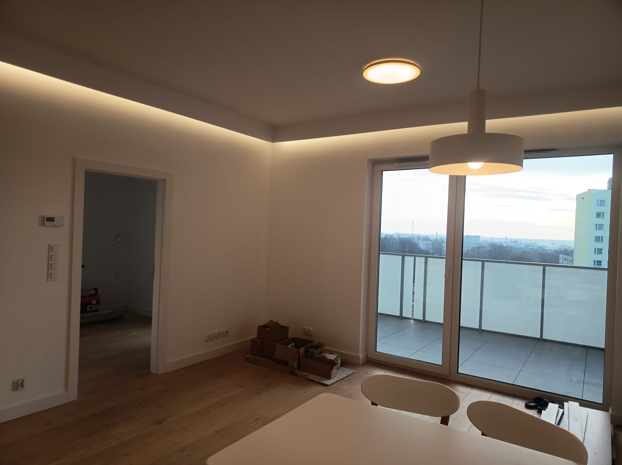 Jasne wnętrze po remoncie: parkiet, białe ściany z oświetleniem LED, balkon z widokiem na miasto. Częściowo widoczne meble i kartony po przeprowadzce.