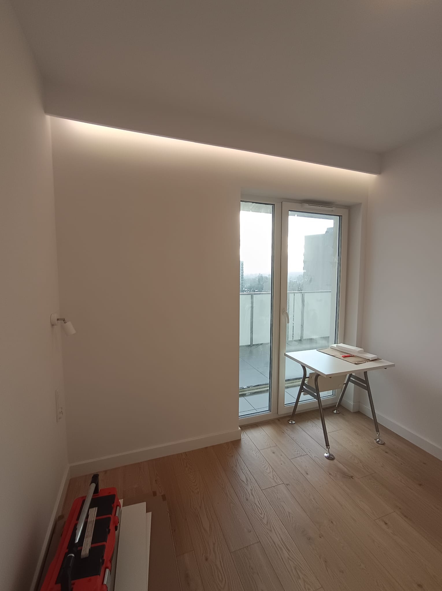 Jasne wnętrze po remoncie: drewniana podłoga, białe ściany z oświetleniem LED, okno balkonowe i narzędzia do układania płytek w rogu. Minimalistyczny design.