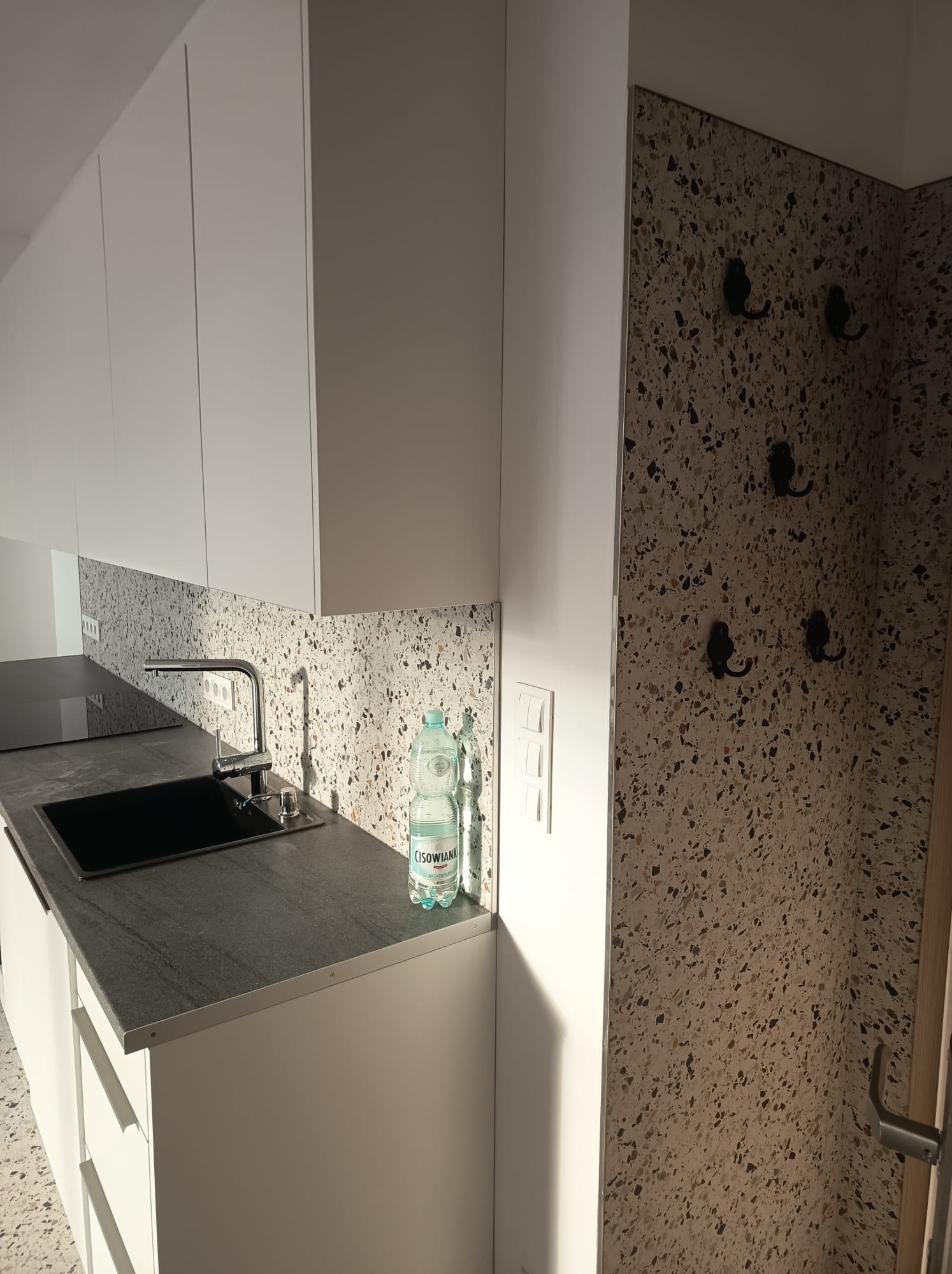 Nowoczesna kuchnia z białymi szafkami, blatem z ciemnego kamienia i mozaikową ścianą. Bateria kuchenna i butelka wody na blacie. Haczyki na ścianie w tle.