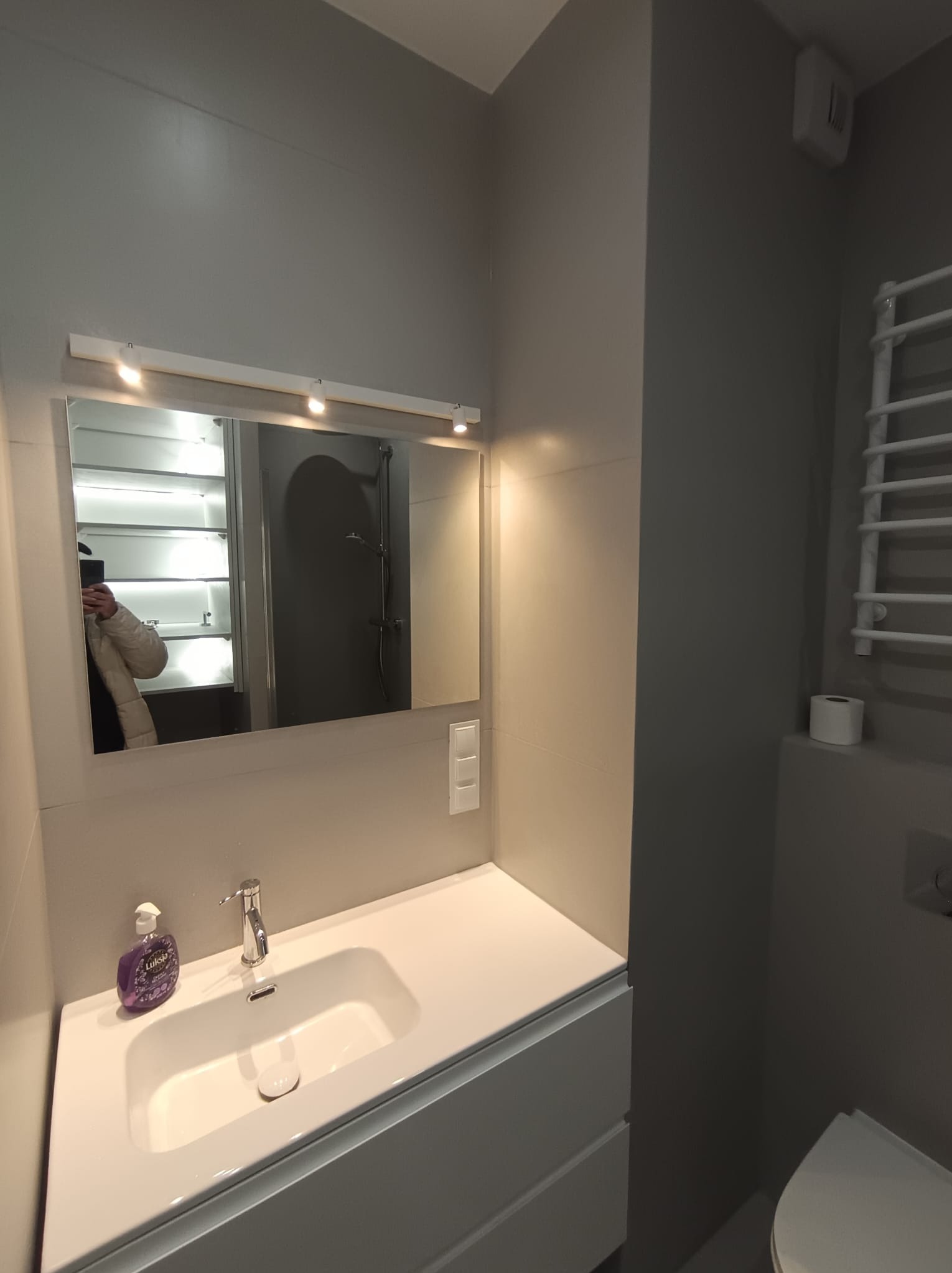 Nowoczesna łazienka w szarościach: biała umywalka z szafką, lustro z oświetleniem LED, grzejnik drabinkowy. Widoczne detale: dozownik mydła i toaleta.