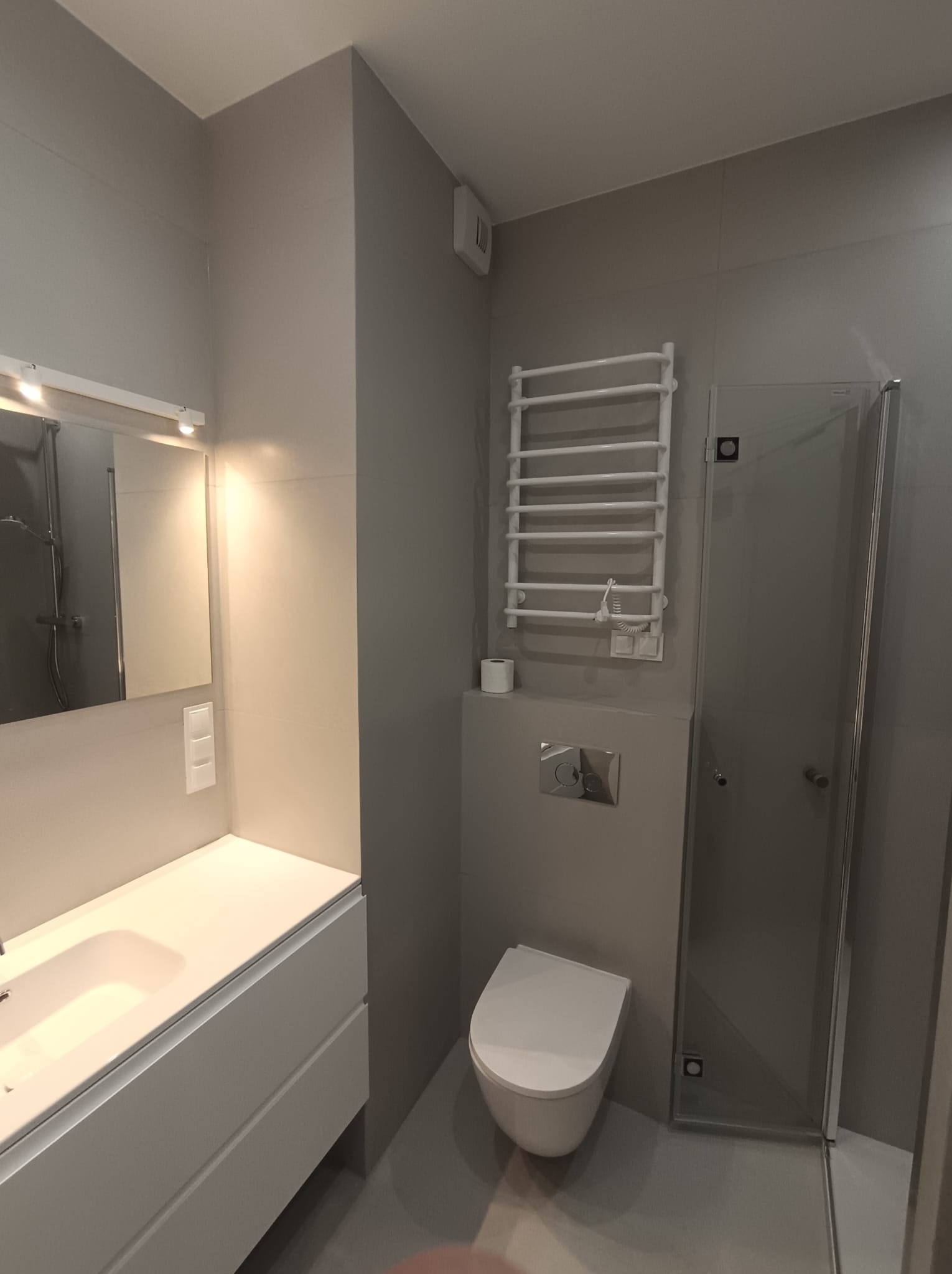 Nowoczesna łazienka w odcieniach szarości z białymi elementami: umywalka z szafką, toaleta podwieszana, grzejnik drabinkowy i kabina prysznicowa.