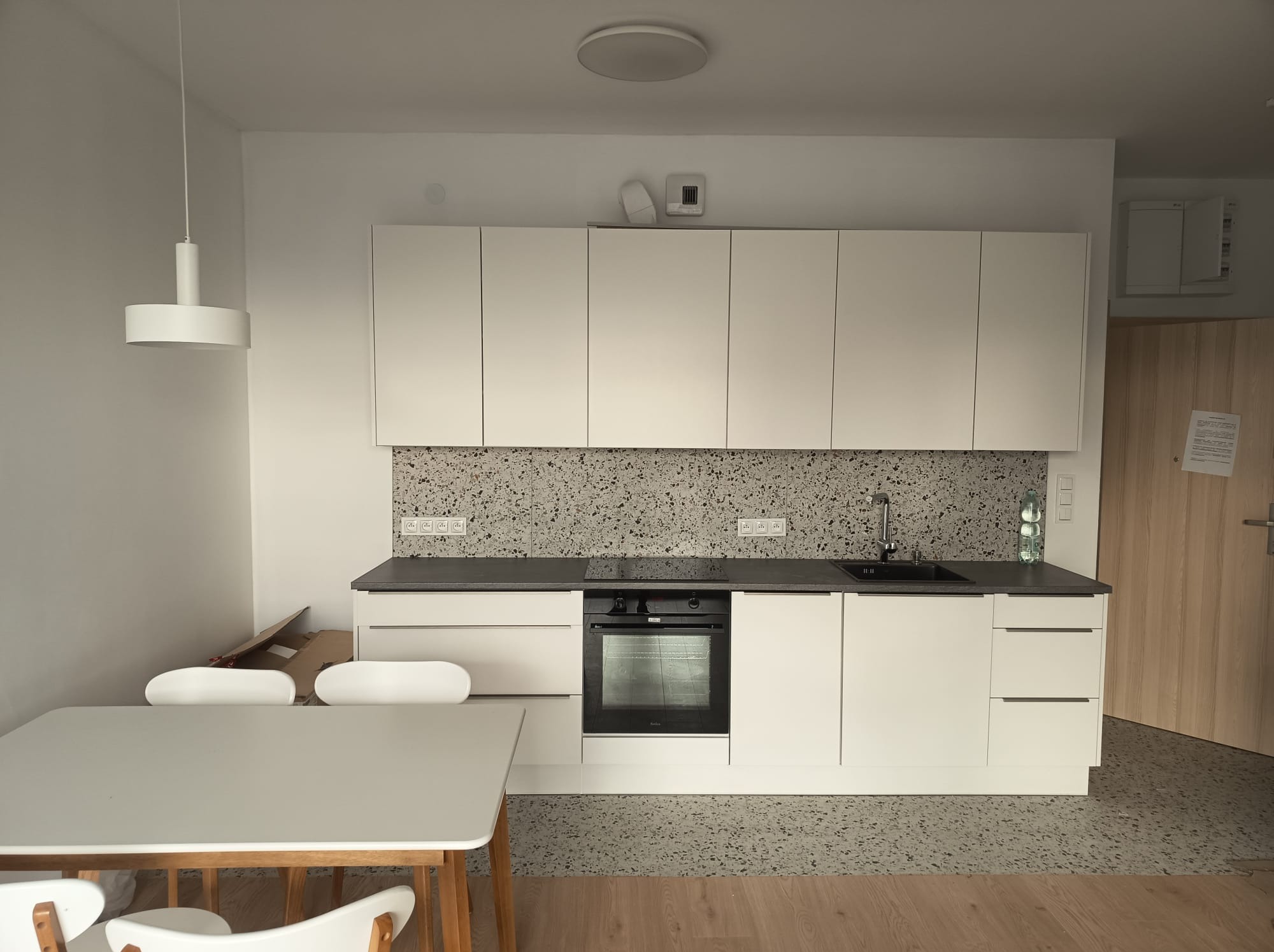 Nowoczesna, minimalistyczna kuchnia z białymi szafkami, szarym blatem i mozaiką na ścianie. Widoczny stół z krzesłami i lampa wisząca. Uwagę zwraca wzór lastryko na podłodze.