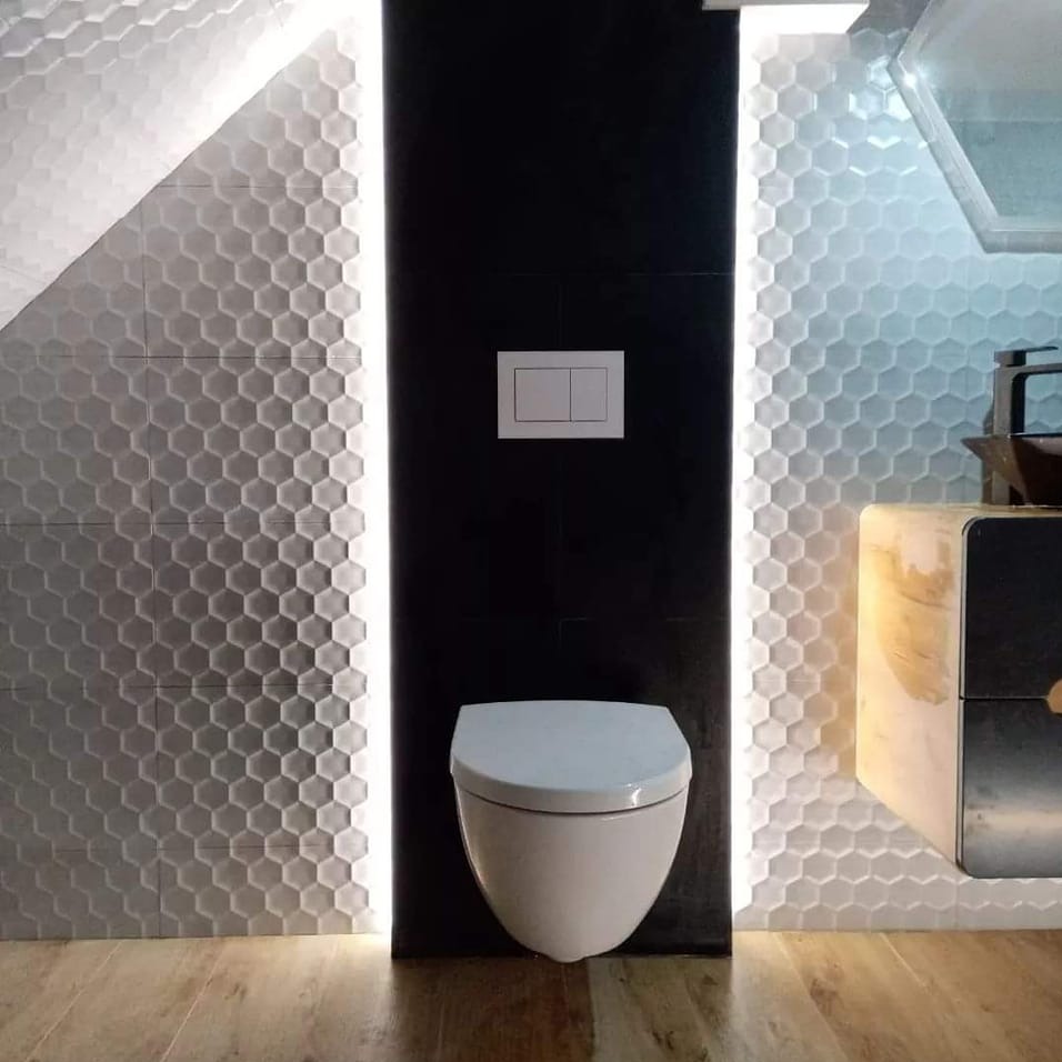 Nowoczesna łazienka z podświetlanymi heksagonalnymi płytkami, wiszącą toaletą i czarną ścianą. Minimalistyczny design z drewnianą podłogą i elementami armatury.