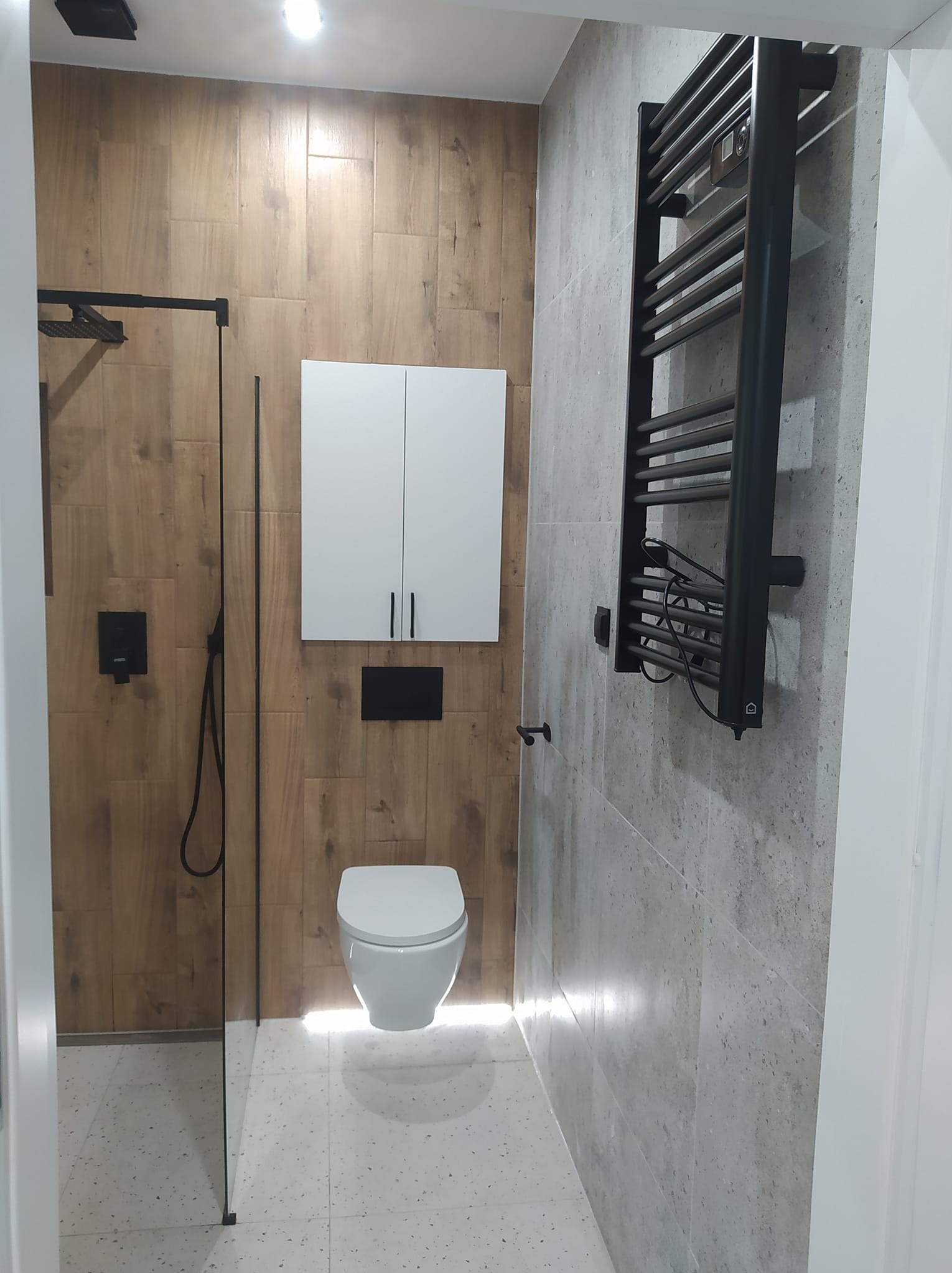 Nowoczesna łazienka po remoncie: kabina prysznicowa ze szklaną ścianką, podwieszana toaleta z podświetleniem LED, czarny grzejnik drabinkowy i drewnopodobne płytki.