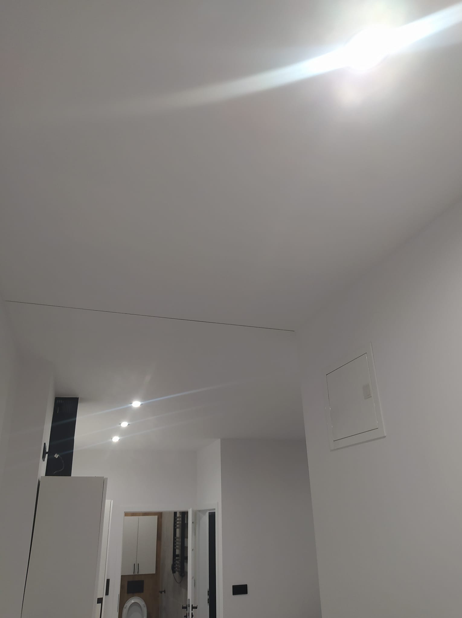 Białe wnętrze łazienki po remoncie, widok z góry, z widocznym oświetleniem sufitowym i szafkami w tle. Minimalistyczny design, świeżo pomalowane ściany.