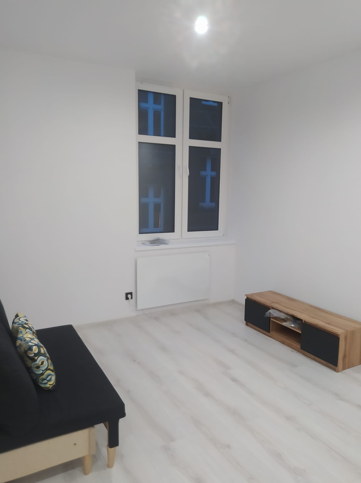 Jasne wnętrze po remoncie: białe ściany, panele podłogowe, okno z niebieską folią ochronną, grzejnik, sofa i szafka RTV. Minimalistyczny design.