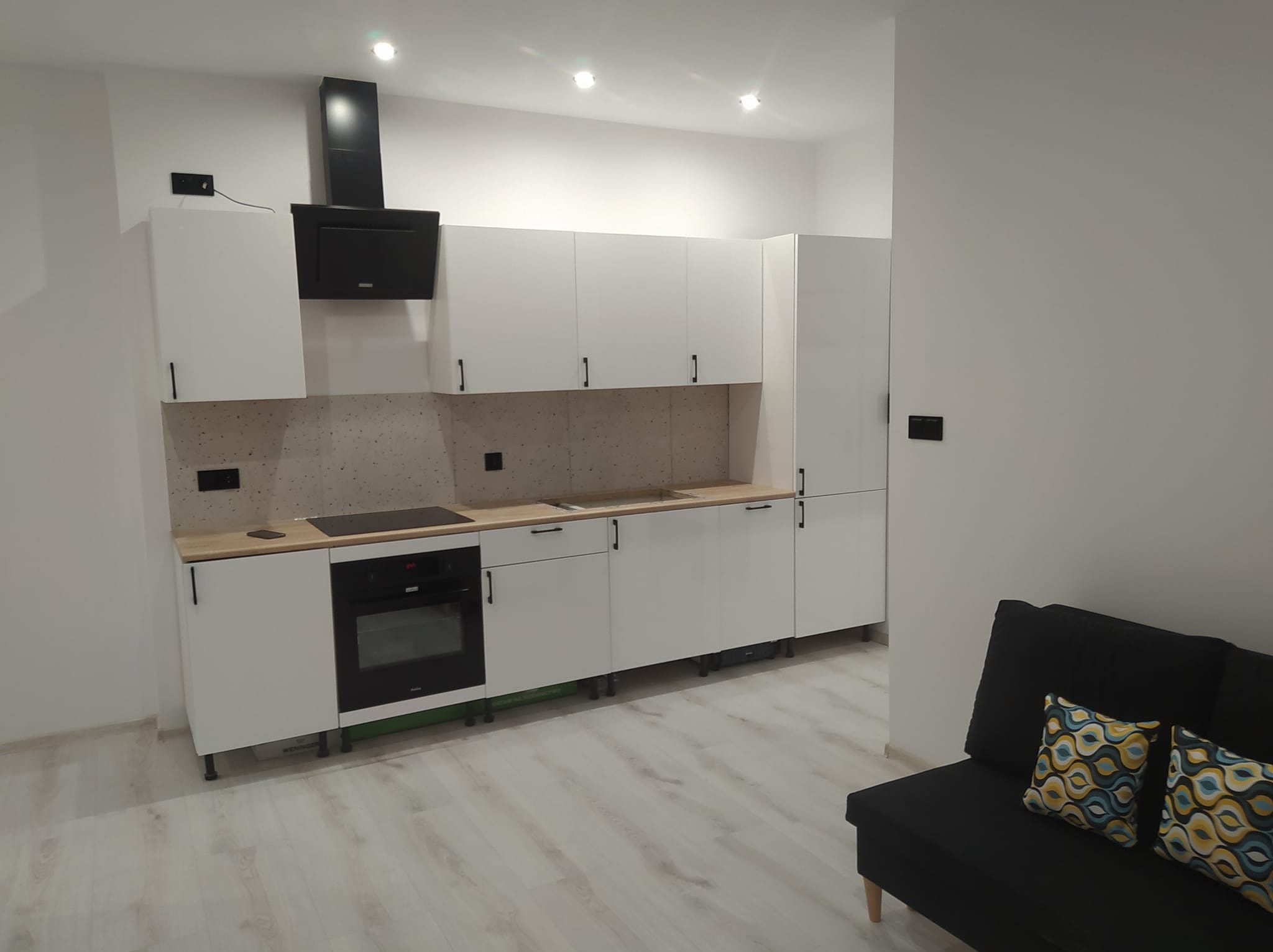 Nowoczesna, minimalistyczna kuchnia z białymi szafkami i drewnianym blatem. Czarny okap i piekarnik kontrastują z jasnym wnętrzem. W tle widoczna sofa.