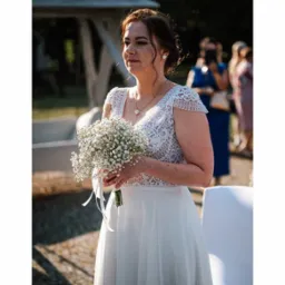 Panna młoda w koronkowej sukni trzyma bukiet gipsówki. W tle goście weselni i biała altana. Delikatne światło podkreśla uroczysty charakter wydarzenia.