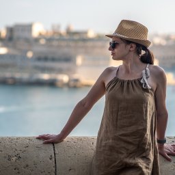 Adrian Cichy - Kobieta w brązowej sukience i słomkowym kapeluszu, oparta o kamienny mur z widokiem na miasto i morze w tle, w okularach przeciwsłonecznych.
