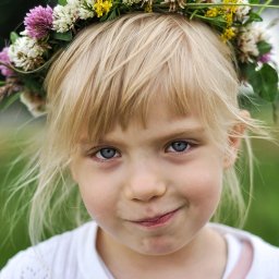 Adrian Cichy - Portret uśmiechniętej dziewczynki z blond włosami i niebieskimi oczami, w wianku z polnych kwiatów na głowie, ubrana w biały sweter