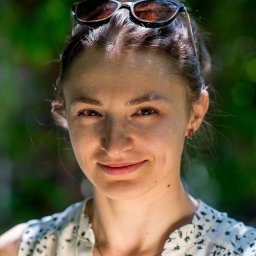 Adrian Cichy - Portret uśmiechniętej kobiety z okularami na głowie, na tle rozmytej zieleni, w bluzce w drobne kwiatki i z naszyjnikiem w kształcie serca.