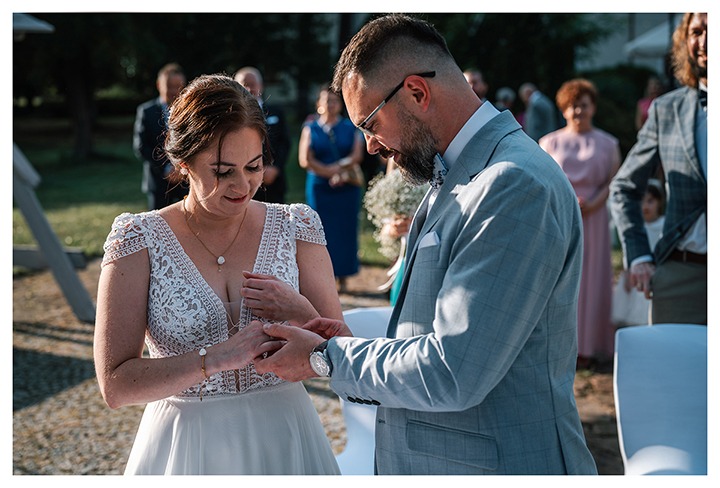 Panna młoda w koronkowej sukni i pan młody w szarej marynarce wymieniają się obrączkami podczas ceremonii ślubnej w plenerze, z gośćmi w tle.